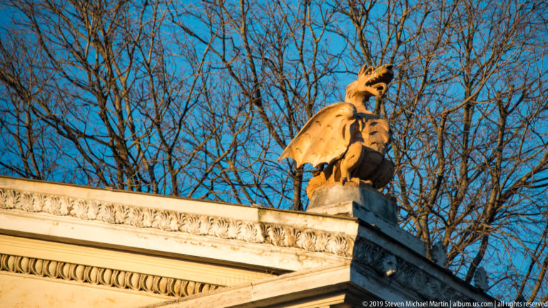 Winged stone dragon perched above a carved classical cornice against leafless branches and blue sky.