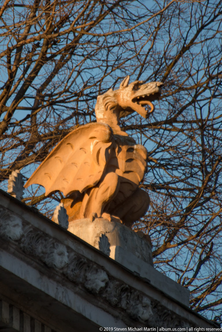 Stone winged dragon with open jaws and folded wings perched on a classical gate beneath bare tree branches.