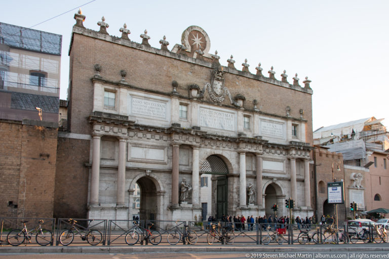 Outer façade of Porta del Popolo with monumental central arch, classical columns, pediment, and sculptural architectural details.