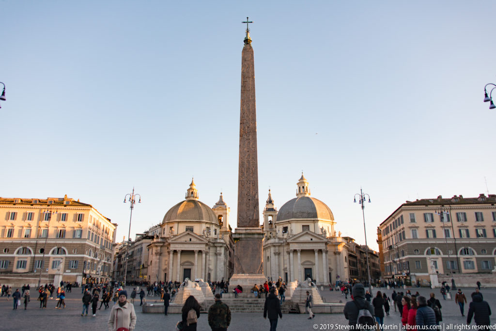 Wide Piazza del Popolo view with central obelisk, twin domed churches, surrounding buildings, and pedestrians.