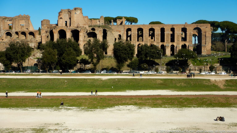 Long arcaded brick ruins on the Palatine Hill above the open Circus Maximus with scattered pedestrians.