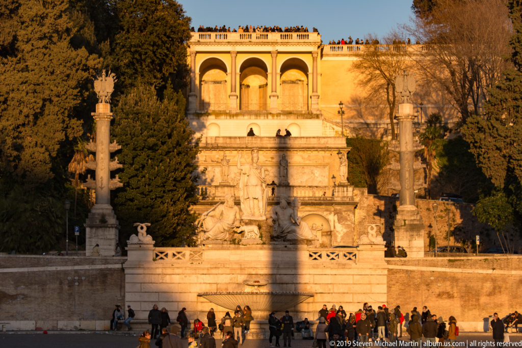 Wide view of the Goddess Rome fountain below trees, terraces, and monumental columns at the edge of Piazza del Popolo.