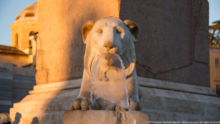 Marble lion fountain and travertine basin at the stepped base of the Flaminio Obelisk in Piazza del Popolo.