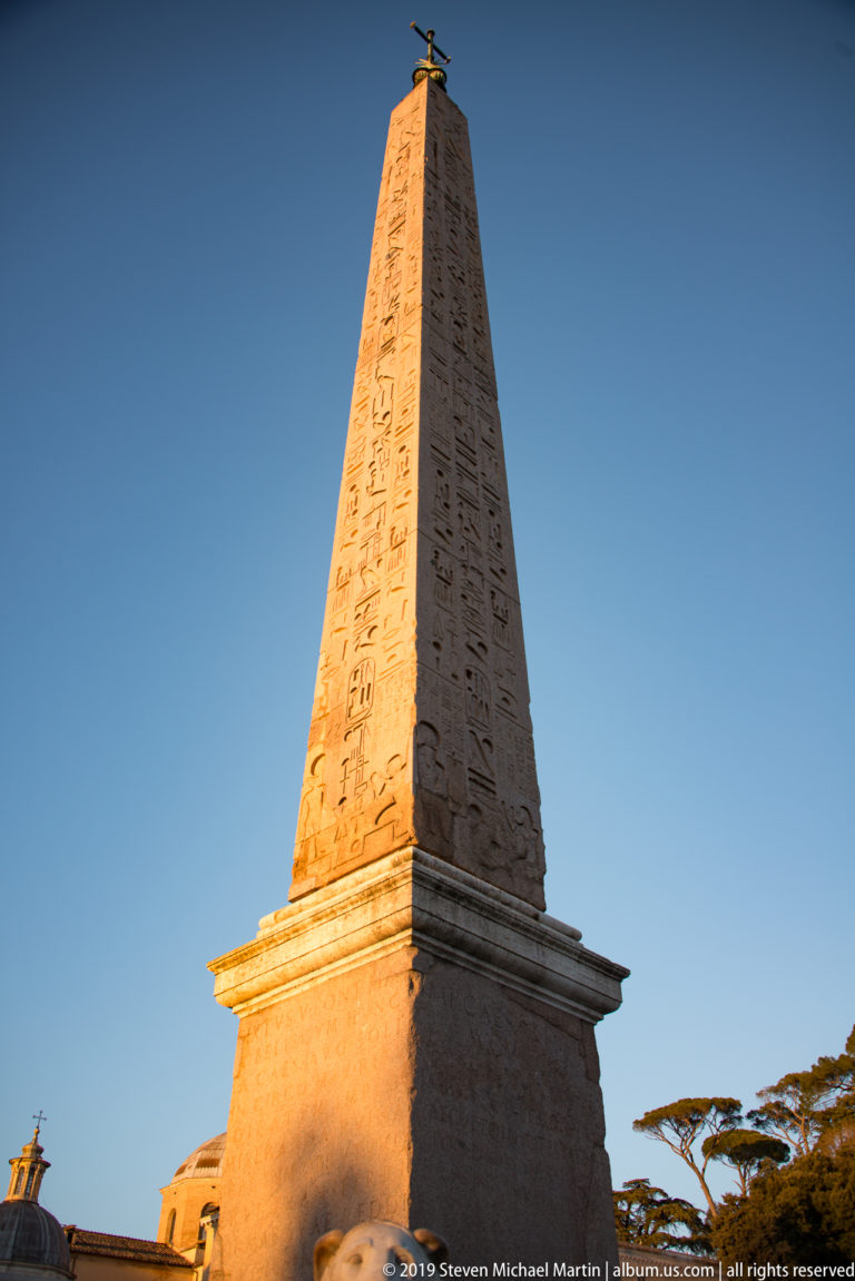 Upper Flaminio Obelisk with carved Egyptian hieroglyphs and a small cross silhouetted against clear blue sky.