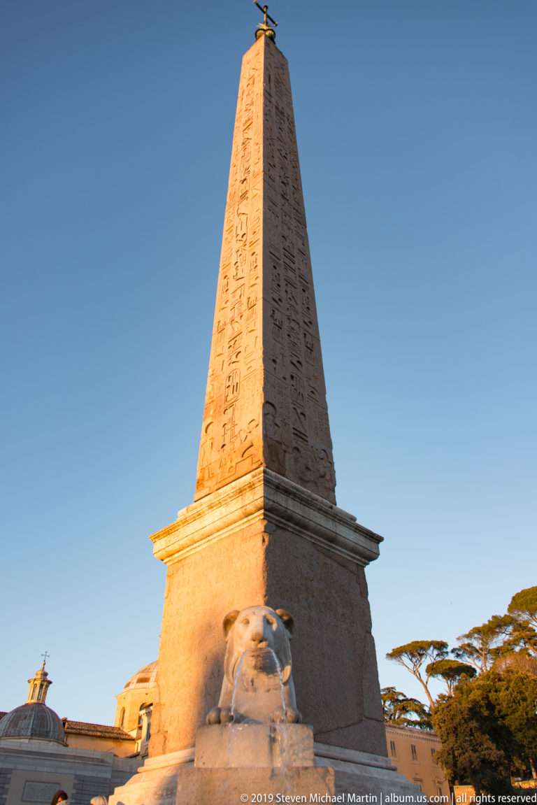Hieroglyph-covered Flaminio Obelisk rising above a marble lion fountain in Piazza del Popolo under blue sky.
