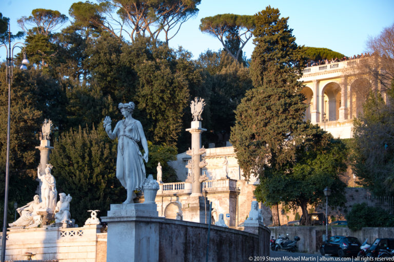 Marble classical statue before fountains, balustrades, trees, and an arched terrace structure at Piazza del Popolo.