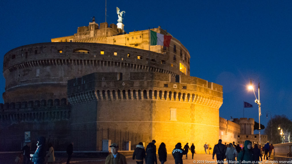 Floodlit Castel Sant’Angelo at night, with cylindrical masonry, Italian flag, rooftop angel, and pedestrians in the foreground.