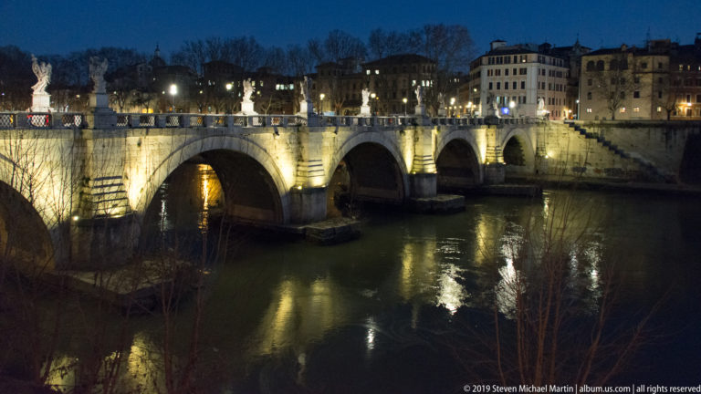 Ponte Sant’Angelo illuminated at night, with stone arches, statues along the balustrade, and golden reflections on the Tiber.