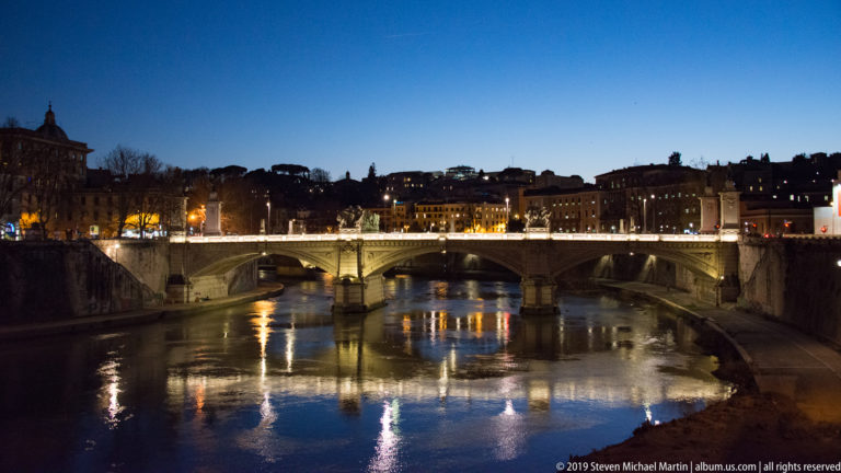Illuminated Ponte Vittorio Emanuele II crossing the Tiber at blue hour, with bridge lights reflected in the river.