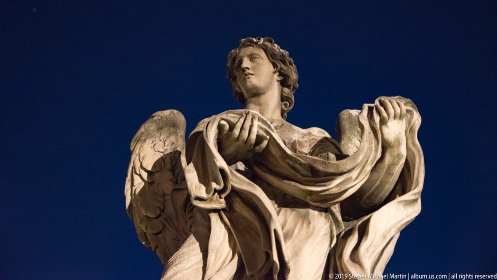 Low-angle night view of a marble angel statue on Ponte Sant’Angelo, with wings and flowing drapery against a dark blue sky.