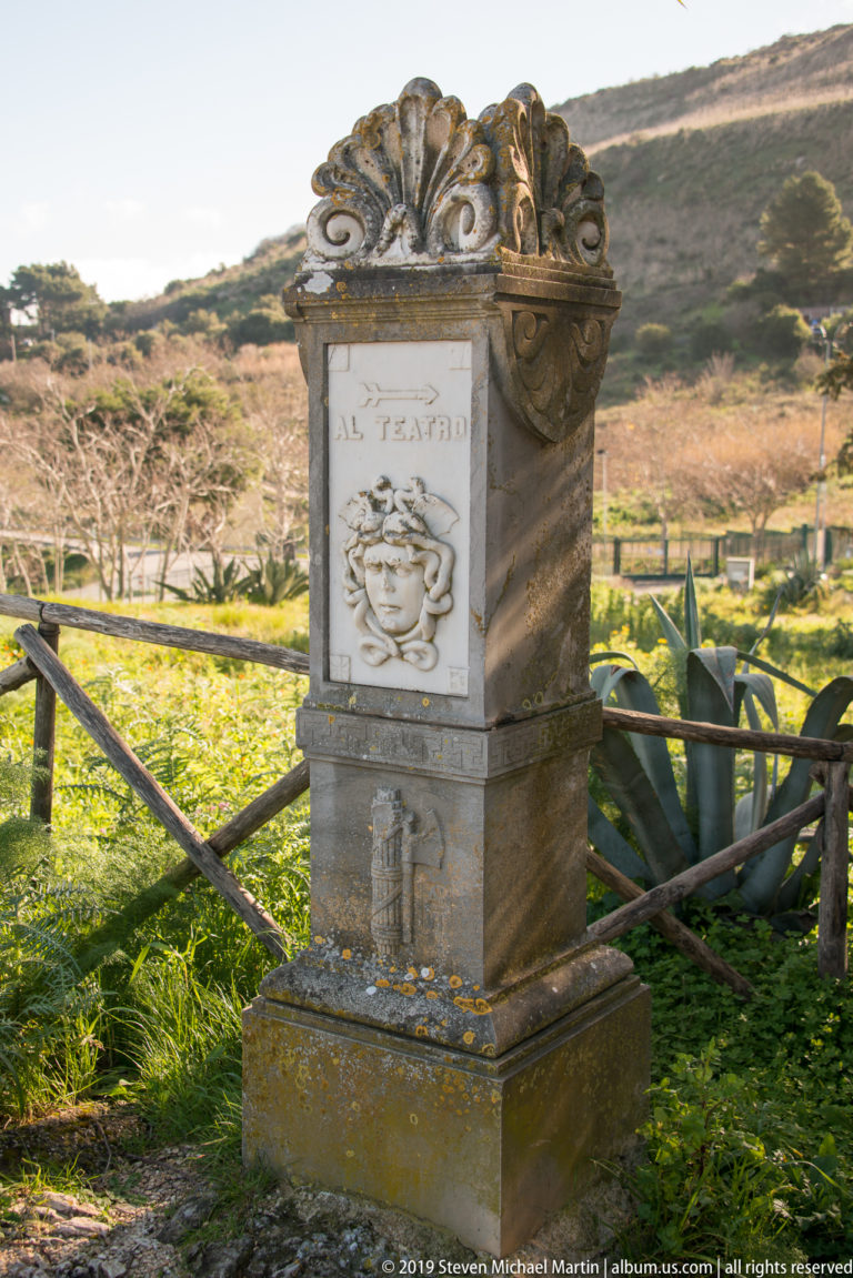 Weathered stone pedestal with a carved classical-style face and ornate crest beside a wooden railing at Segesta.