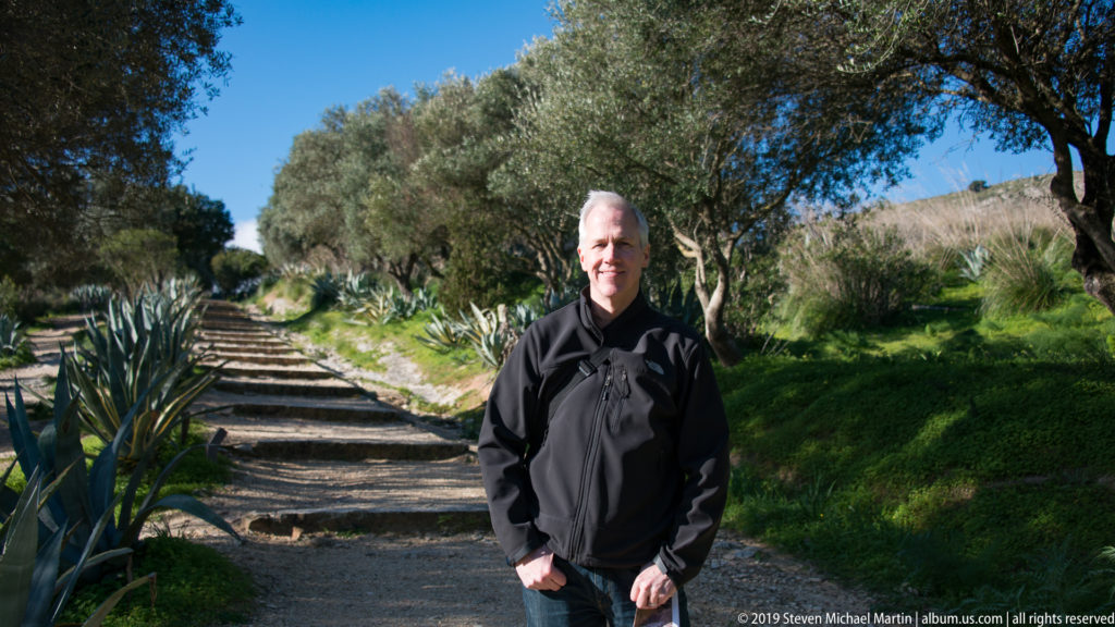 Visitor standing on a stepped gravel path lined with olive trees and agaves at Segesta.