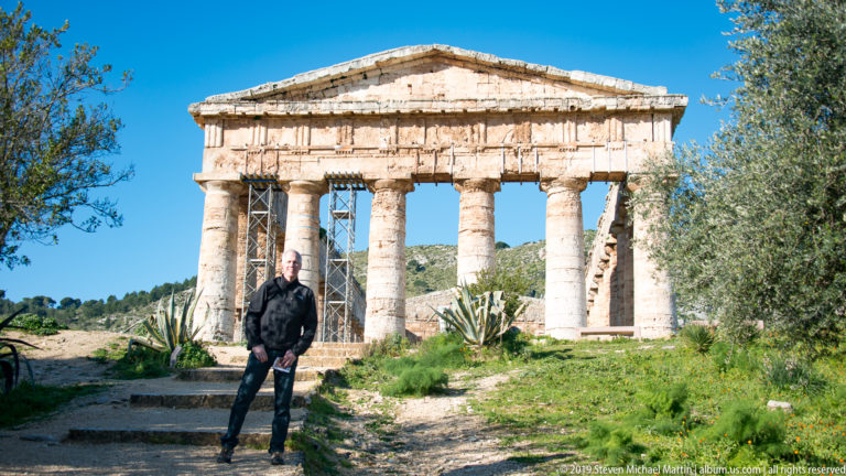 Visitor standing on a path before the Doric façade of the Temple of Segesta, framed by trees and agaves.