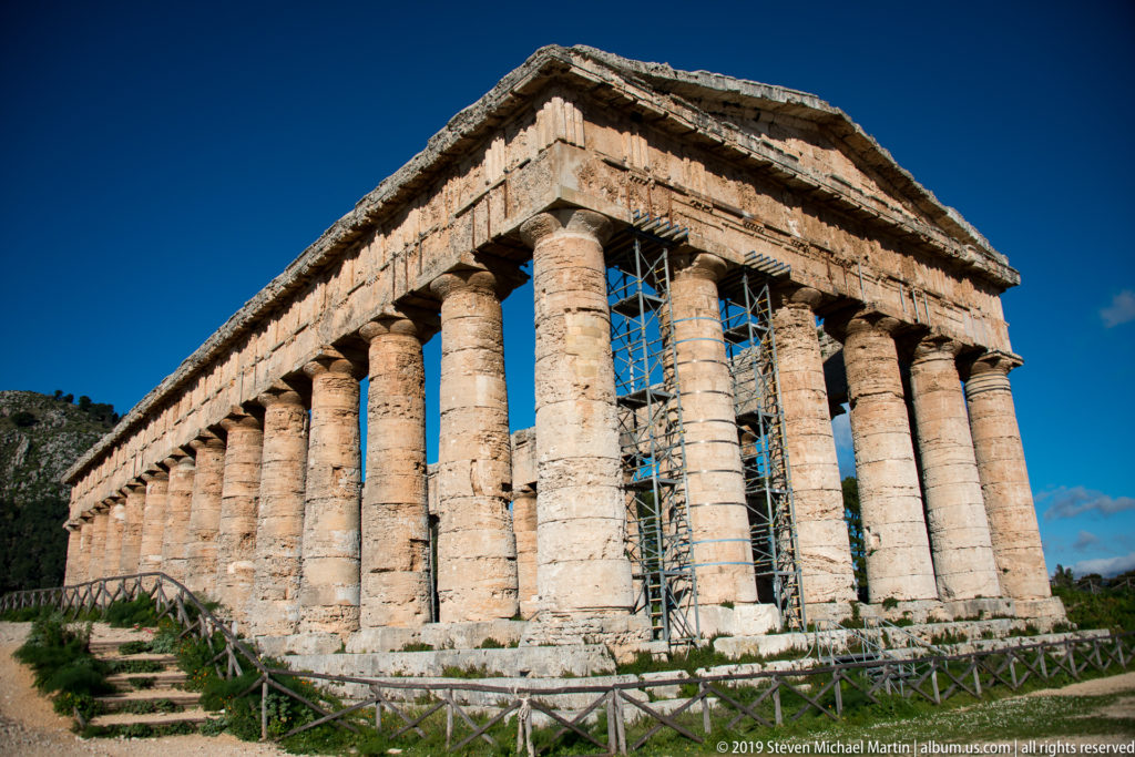 Three-quarter exterior view of the Temple of Segesta with Doric columns, stone steps, railing, and conservation scaffolding.