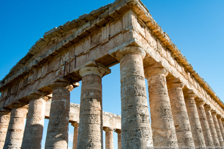 Close upward view of weathered Doric columns and the stone entablature at a corner of the Temple of Segesta.