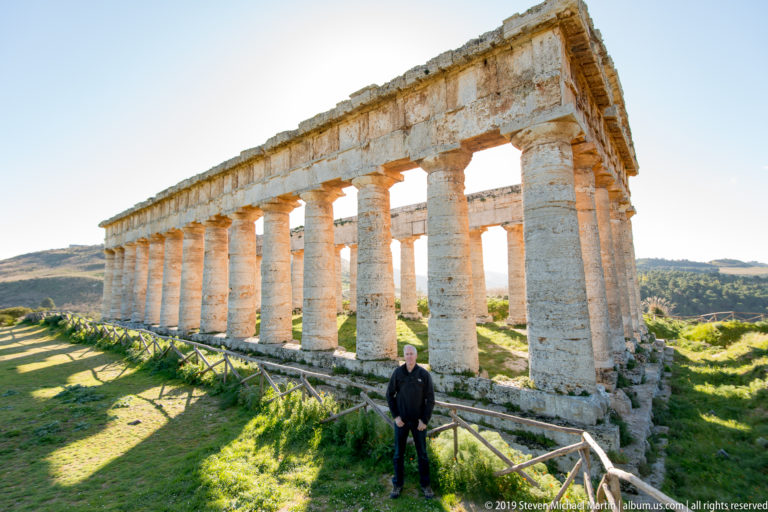 Long sunlit colonnade of the Temple of Segesta with a visitor standing beside the wooden perimeter railing.
