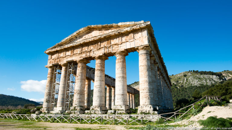Low oblique view of Segesta’s Doric temple with façade columns, receding side, wooden rail, and rocky hill.