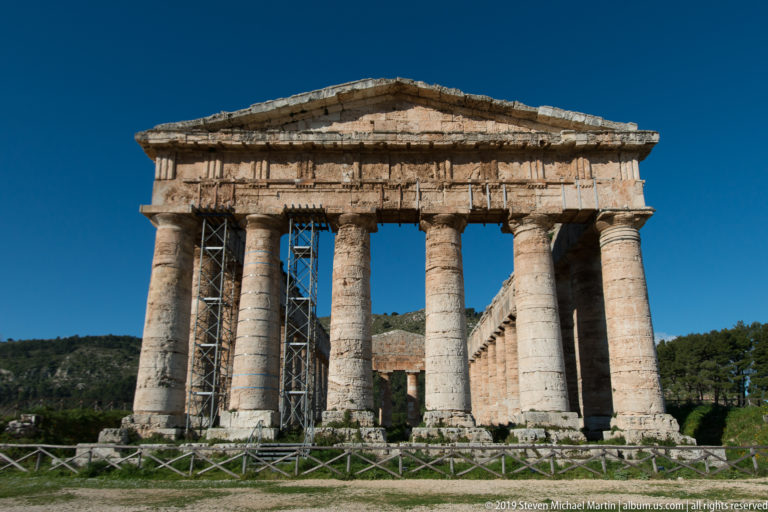 Centered front view of Segesta’s Doric temple with six columns, triangular pediment, scaffolding, and low barrier.