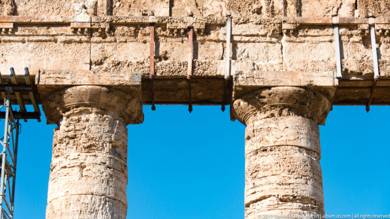 Close view of two weathered Doric capitals supporting the temple entablature with metal conservation elements above.