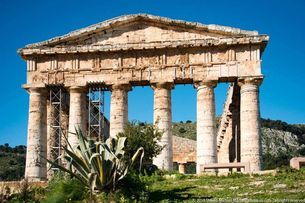 Front of Segesta’s Doric temple with six columns, triangular pediment, interior scaffolding, and an agave-like plant.