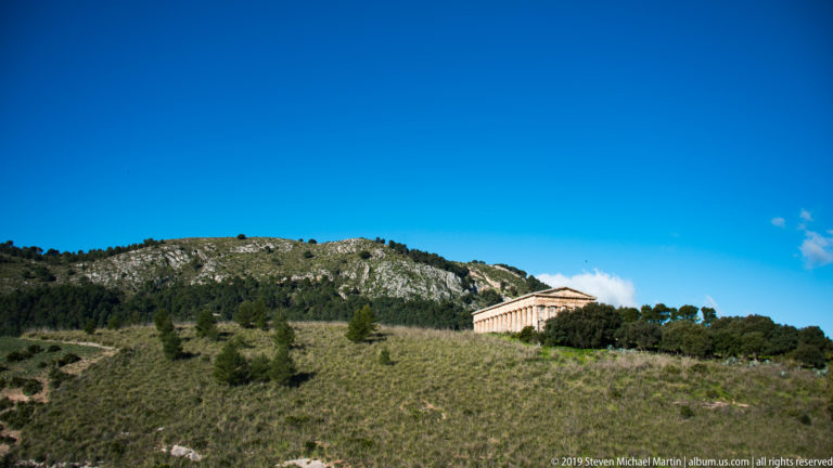 Distant side view of Segesta’s Doric temple above trees on a grassy hillside under deep blue sky.