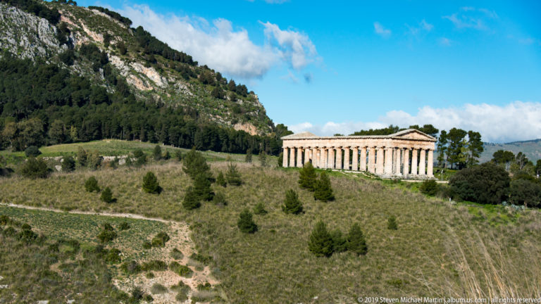 Long side of Segesta’s Doric temple extending across a grassy hillside beneath a wooded rocky ridge.