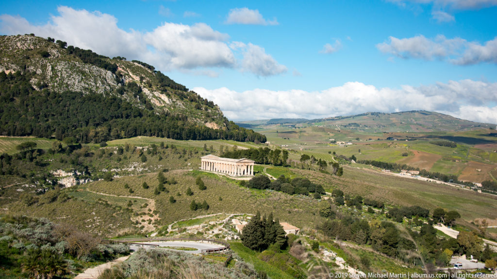 Distant Doric temple at Segesta on a green slope beneath a wooded rocky ridge.
