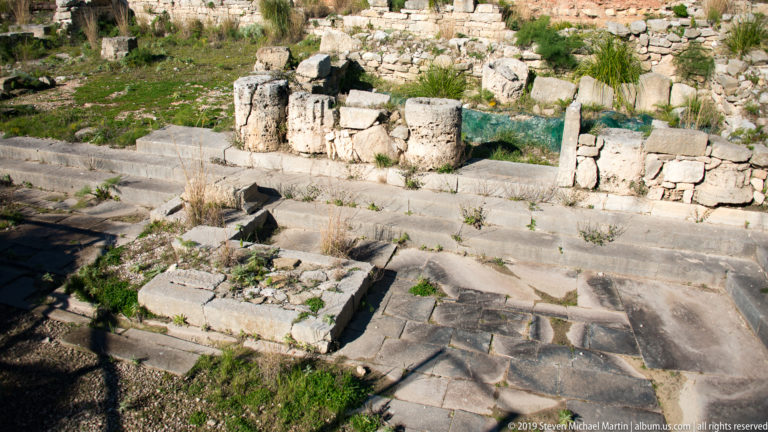 Excavated stone remains with column fragments, steps, paving, and low walls at Segesta.
