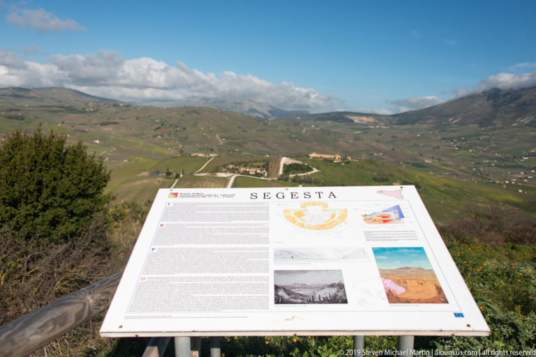 SEGESTA interpretive panel in the foreground overlooking rolling fields, roads, and mountain ridges.