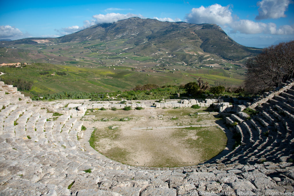 Circular orchestra and curved stone seating of Segesta’s theater with green mountains rising beyond.