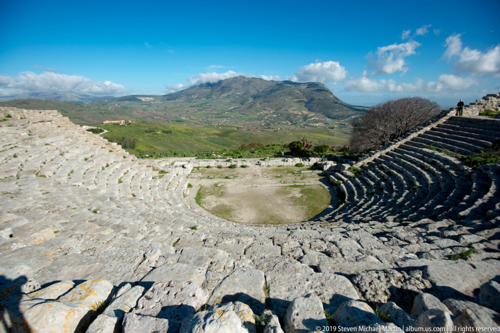 Wide view over Segesta’s curved theater seating and orchestra toward green fields and mountain ridges.
