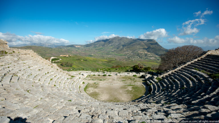 Ancient stone theater descending toward its orchestra with a broad green mountain valley beyond.
