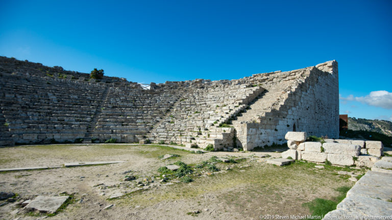 Curved theater seating meeting a massive pale-stone side wall with steps rising along its inner face.