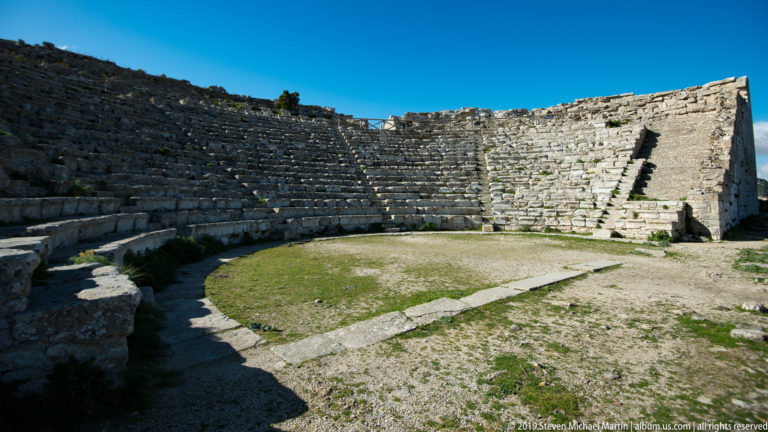 Empty orchestra and curving stone seating inside Segesta’s ancient theater beneath a clear blue sky.
