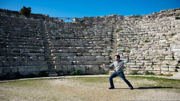 Visitor standing in the orchestra before curving tiers of ancient stone theater seating at Segesta.