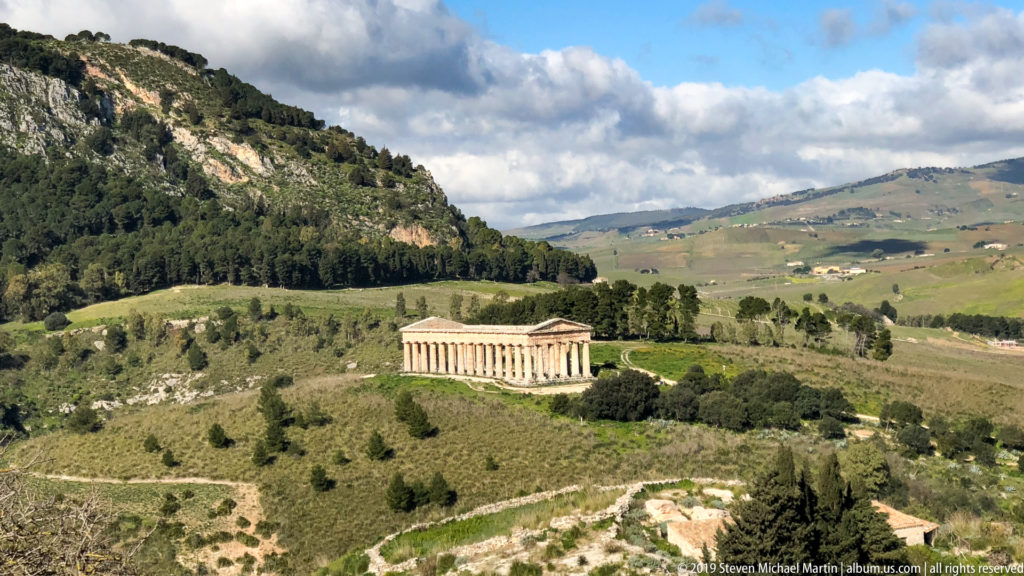 Doric temple at Segesta on a green ridge amid wooded slopes, fields, and distant mountains.