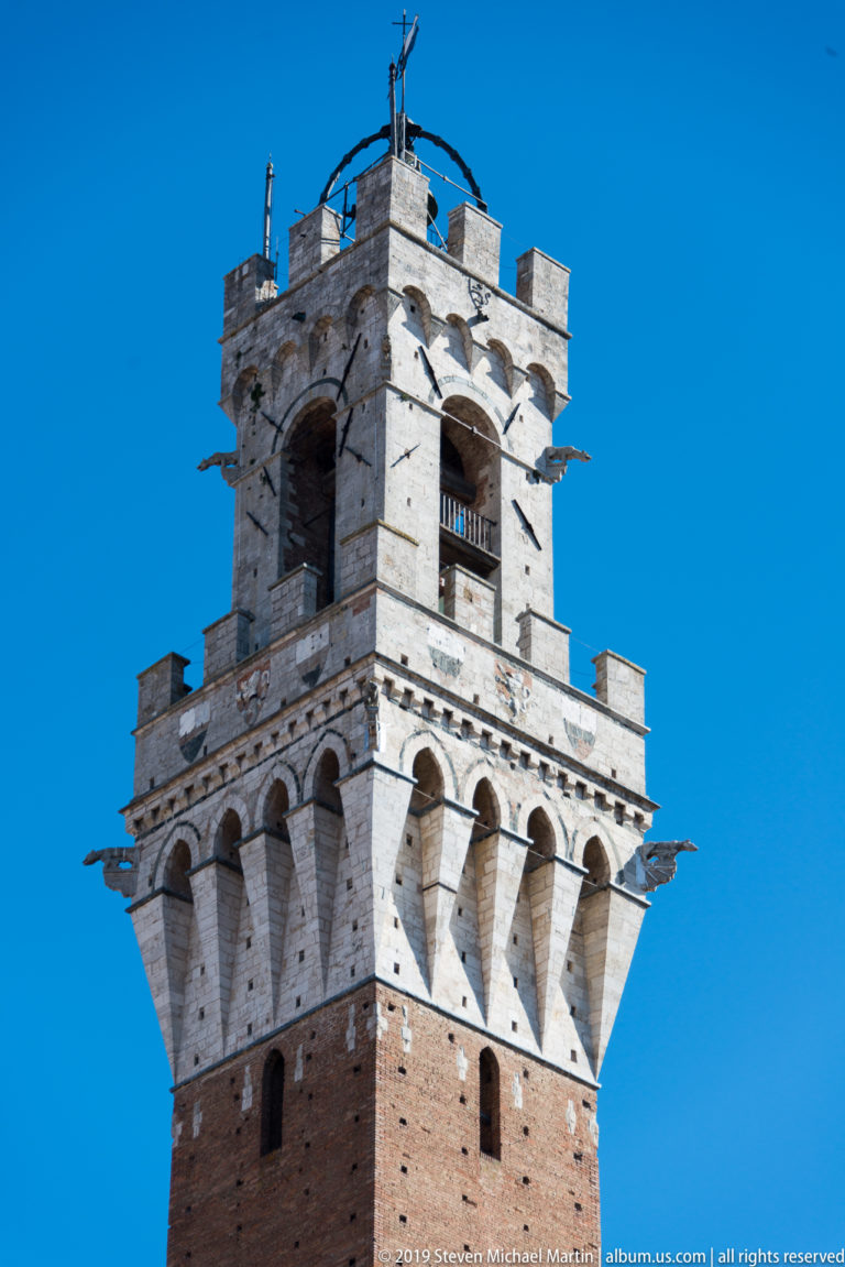 Close upward view of Torre del Mangia’s pale stone belfry, bell opening, projecting corbels, and crenellated crown against blue sky.