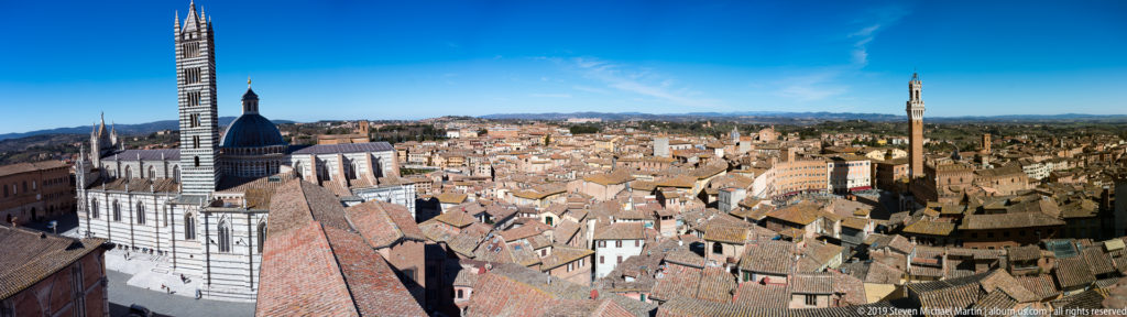 Wide Siena panorama with striped cathedral, dome and campanile at left, dense terracotta roofs, and Torre del Mangia at right.