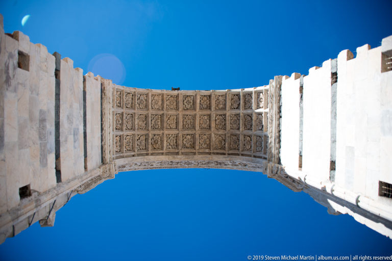 Upward view through a pale marble arch with square carved coffers and rosettes framed against a clear blue sky.