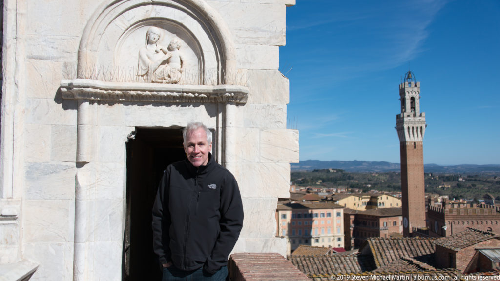 Man in black standing in a small carved stone doorway high above Siena, with Torre del Mangia visible in the background.