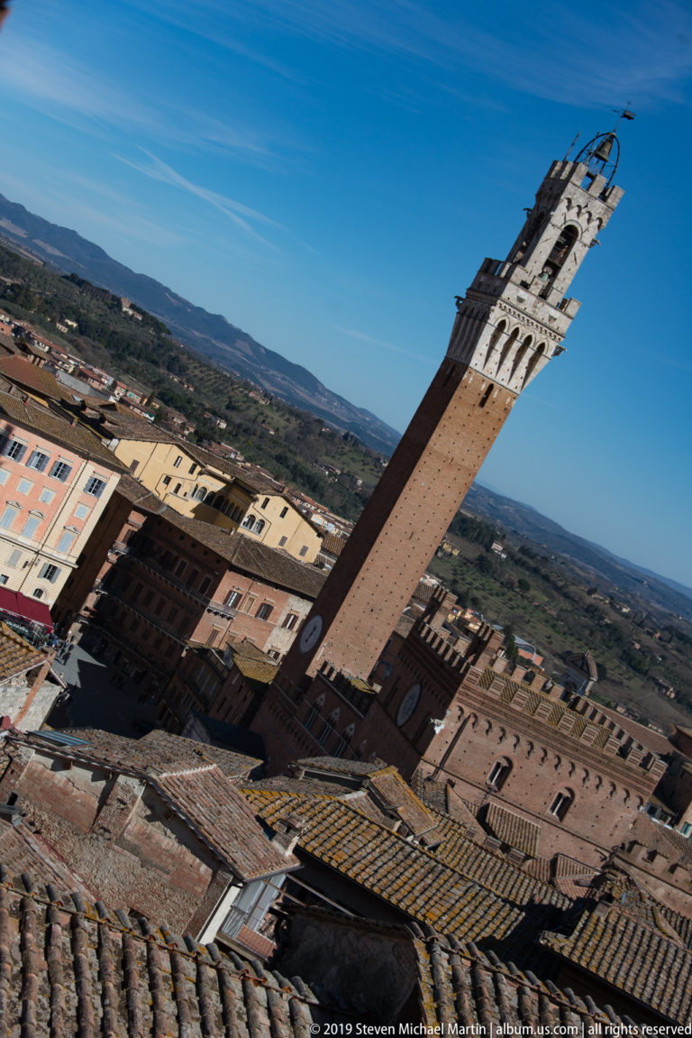 Diagonal high-angle view of the Torre del Mangia rising from Palazzo Pubblico above Siena’s rooftops and distant countryside.