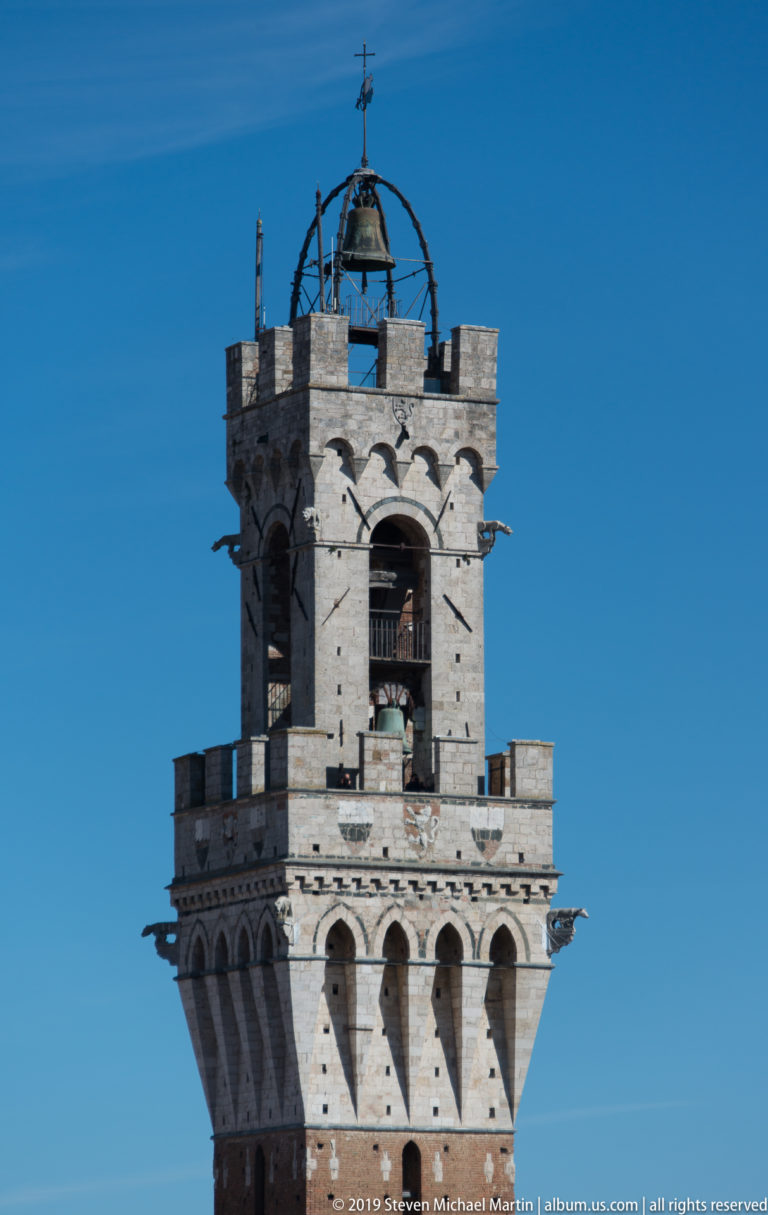 Close view of Torre del Mangia’s pale stone belfry, open bell arches, projecting corbels, and metal crown against blue sky.