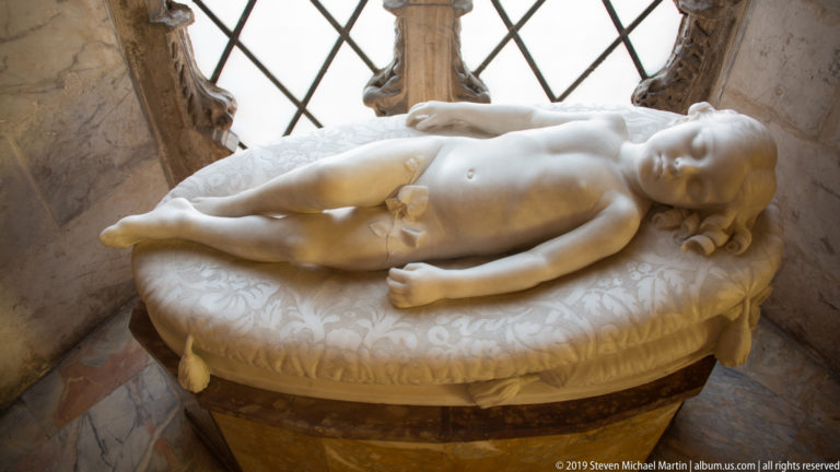 White marble sculpture of a reclining infant resting on a carved cushion beside a latticed architectural opening.