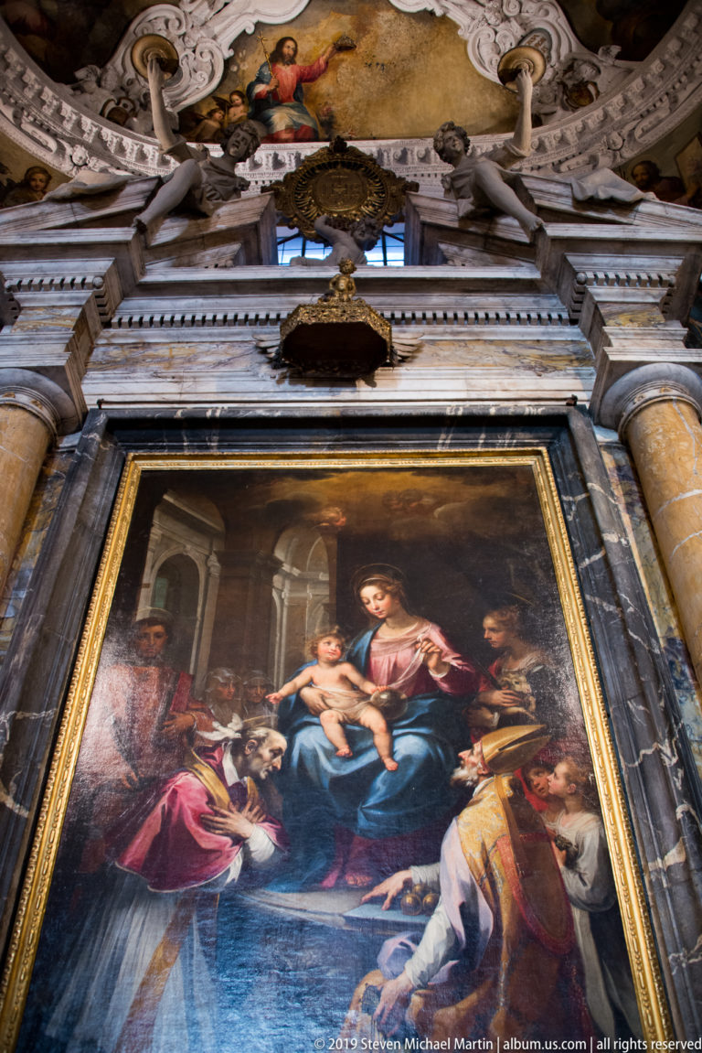 Large Madonna and Child altarpiece with saints in a dark marble frame beneath sculpted and gilded chapel decoration.