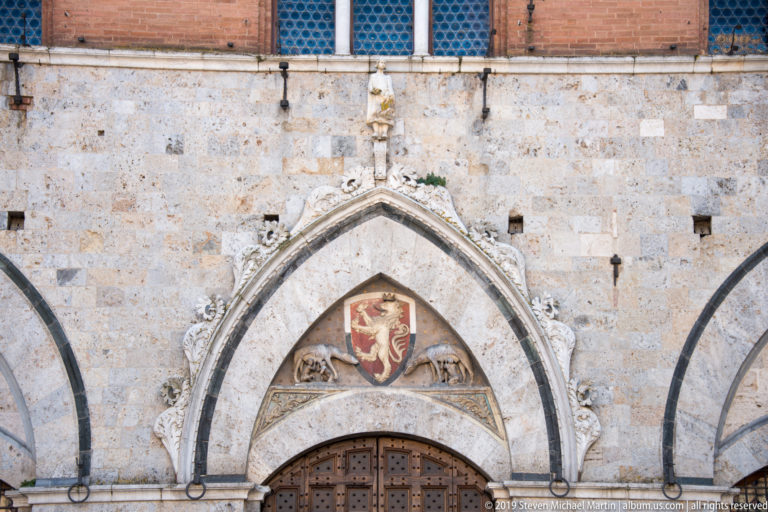 Pointed Gothic doorway with wooden doors, carved pale stone moldings, and a heraldic lion relief in the tympanum.
