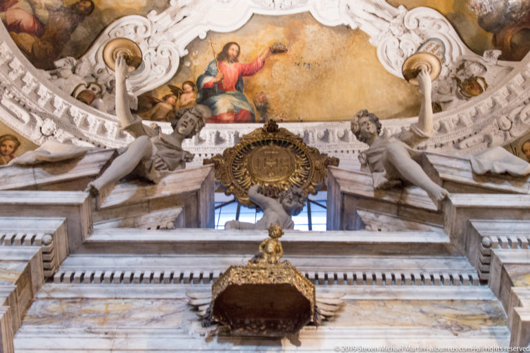 Upward view of gilded altar ornament, marble architecture, reclining stucco figures, circular emblem, and a painted ceiling scene.