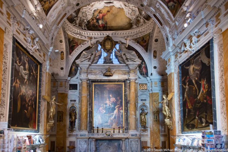 Ornate chapel interior with marble altar, central painting, large side canvases, white stucco sculpture, and painted vault.