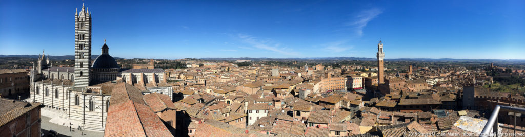 Panoramic Siena skyline with the striped cathedral at left, terracotta roofs across the center, and Torre del Mangia at right.