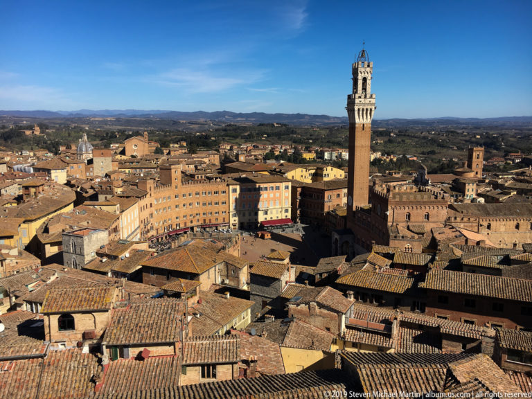 Elevated view over terracotta roofs to Piazza del Campo, Palazzo Pubblico, and the tall Torre del Mangia, with hills beyond.