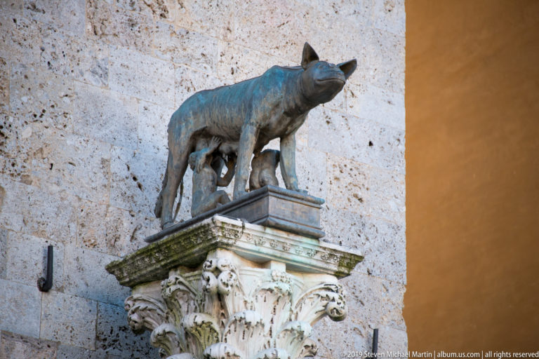 Bronze she-wolf nursing Romulus and Remus atop an ornate carved stone capital projecting from a pale wall.